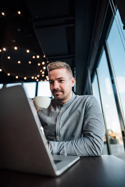 Handsome young entrepreneur looking at laptop screen and drinking coffee. Low angle image.