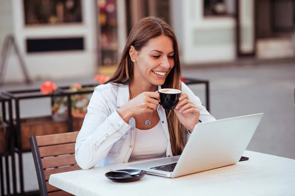 微笑的年轻女子一边看着笔记本电脑 一边在户外咖啡馆喝咖啡 图库照片