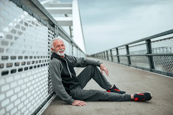 Portrait of a senior sportsman relaxing after a run. Sitting on city bridge, looking at camera.