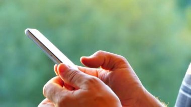 Onlar bir smartphone ellerinde tutmak ve doğa yeşil bir arka plan üzerinde bakmak. Telefonun dokunmatik ekran sayfalarında parmakları başparmak.