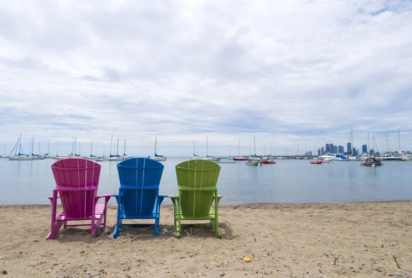 Multicoloured Muskoka Cottage chairs on the sand the beach looking out to the harbor and downtown Etobicoke in the background