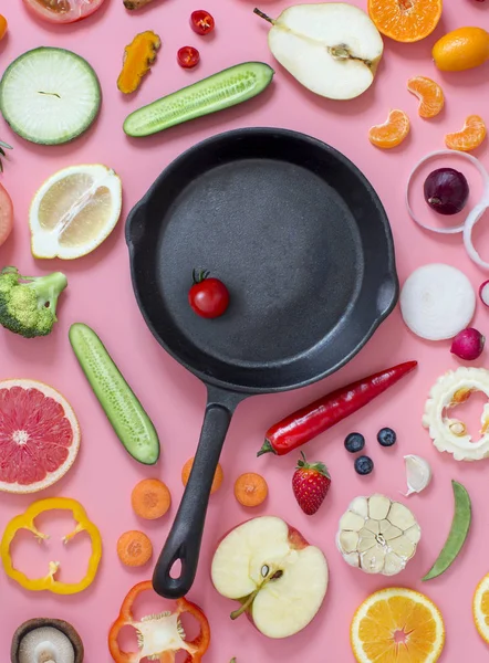 Iron frying pan on colourful sliced vegetables and fruits pink background.