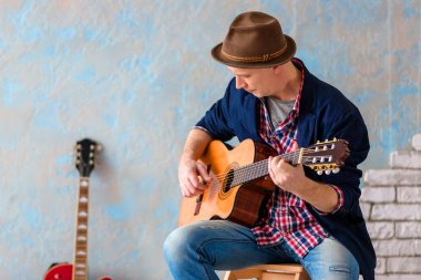 Şapkalı bir müzisyen sandalyeye oturur ve klasik gitar çalar.