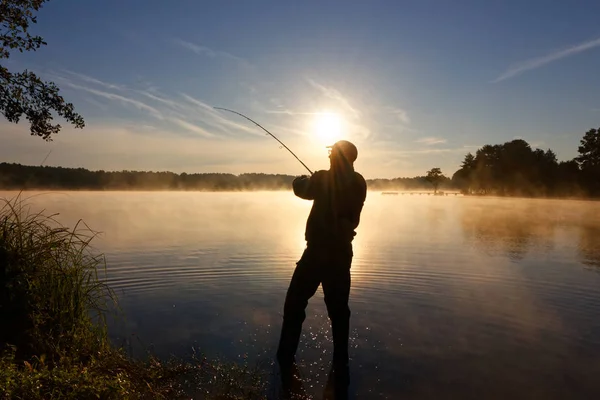 渔夫的剪影在湖和捉住鱼在雾的日出期间 — 图库照片