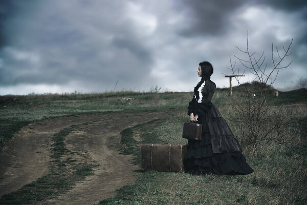 Outdoors portrait of a victorian lady in black sitting alone on the road with her luggage.