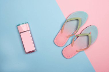 Pembe ve mavi pastel arka planda şık plaj parmak arası terlik ve pembe güneş kremi şişesi, üst görünüm. Kopyalama alanı ile yaz konsepti.