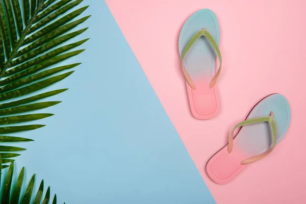 Stylish beach flip-flops on pink and blue pastel background with palm leaf, top view. Summer concept with copy space.