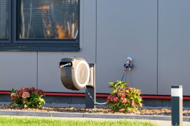 Çağdaş bir binanın önündeki duvara monte edilmiş musluğa bağlı çekilebilir bahçe hortumu makarası çiçek açan ortanca çiçekleriyle. Verimli bahçıvanlık, açık hava su yönetimi kavramı.