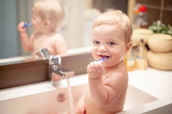 A little sweet little girl with short hair and a white sweater sitting at home in the bathroom on the sink and brushing her teeth with a large blue toothbrush. — Stock Photo, Image
