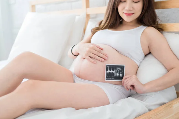 ベッドの上の赤ちゃんの超音波写真を保持している妊娠中の女性 — ストック写真
