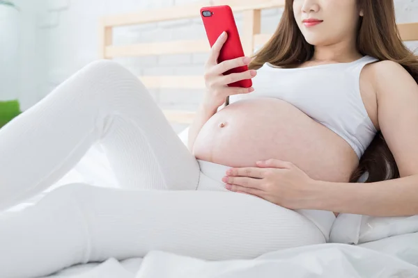 妊娠中の女性がベッドの上に座っていると喜んで電話を使用して — ストック写真