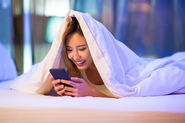 女人高兴地使用智能手机 晚上躺在被子下的床上 — 图库照片