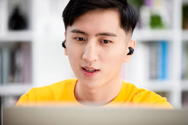 アジア系の若い男性の学生は インターネット上の語学コースや自宅のノートパソコンから情報を検索するために無線イヤホンを使用します — ストック写真