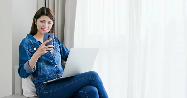 アジアの女性はスマートフォンを見てラップトップを使って家で働いています — ストック写真