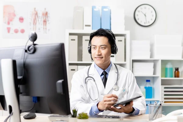 テレメディシンの概念 アジアの男性医師は オンラインデジタルタブレット上で患者と書き込みノートに話すヘッドセットを着用 — ストック写真