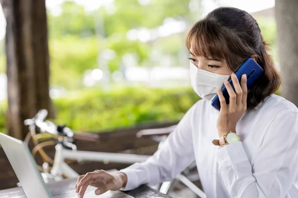 Asian businesswoman wear face mask and talk on phone to work in the park