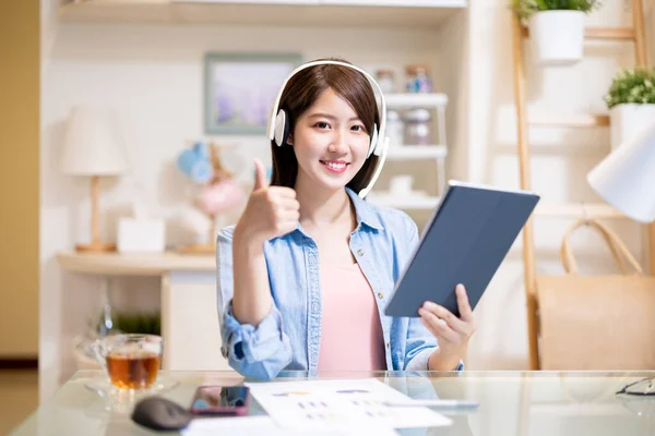 戴着耳机的亚洲女人微笑着看着你 并在家里举着带着大拇指的数码平板电脑 — 图库照片