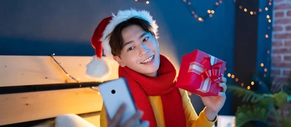 스카프와 크리스마스 모자를 이시아 청년이 스마트폰으로 비디오 채팅을 친구에게 상자를 — 스톡 사진