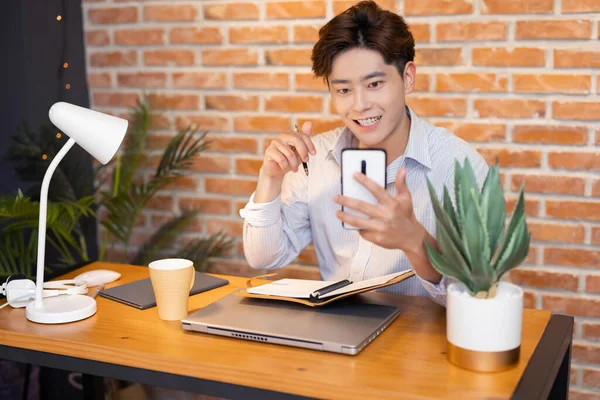 아시아인젊은 사업가는 스마트폰을 사용하여 비디오 회의에 참여하고 집에서 필기를 — 스톡 사진