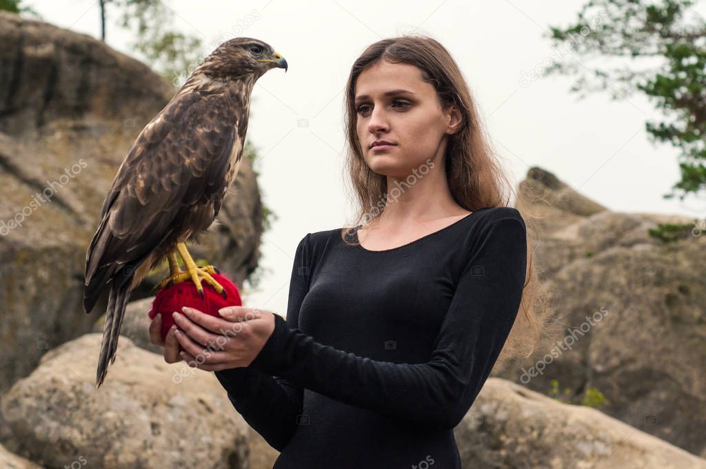  Beautiful girl holding a falcon in her arms. Brunette among the rocks holds a wild bird on a ball of red threads.