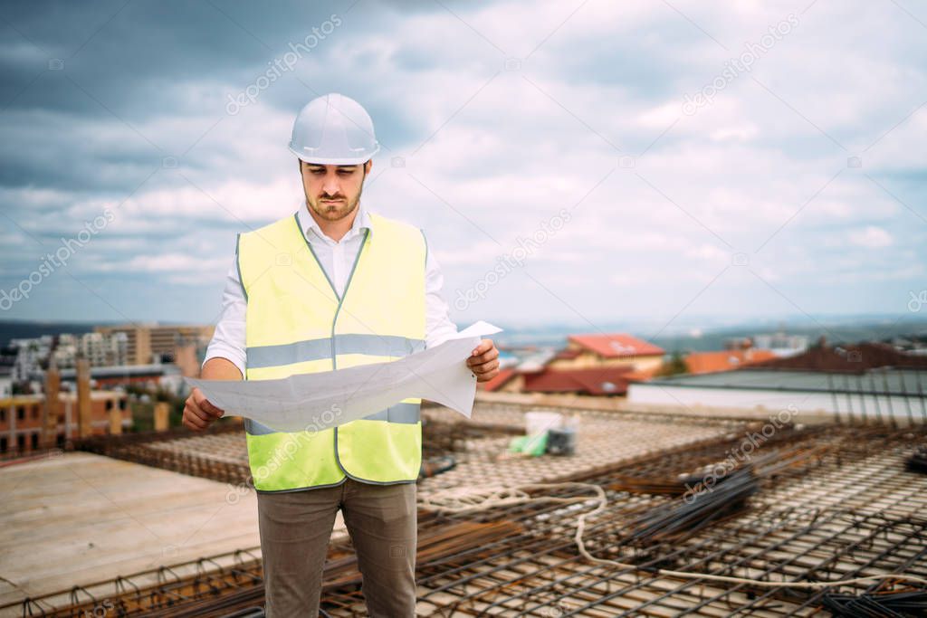 Portrait of male engineer wearing safety equipement on construction site while supervising construction works. Engineer reading blueprints