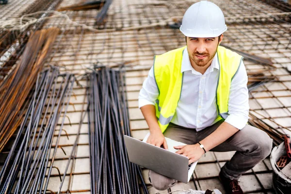 Industrial construction engineer working on laptop, wearing safety equipement and coordinating workers