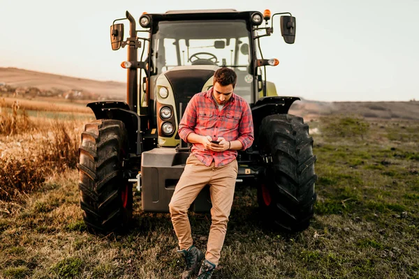 Modern agriculture with technology and machinery concept. Portrait of smiling farmer using smartphone and tractor at harvesting.
