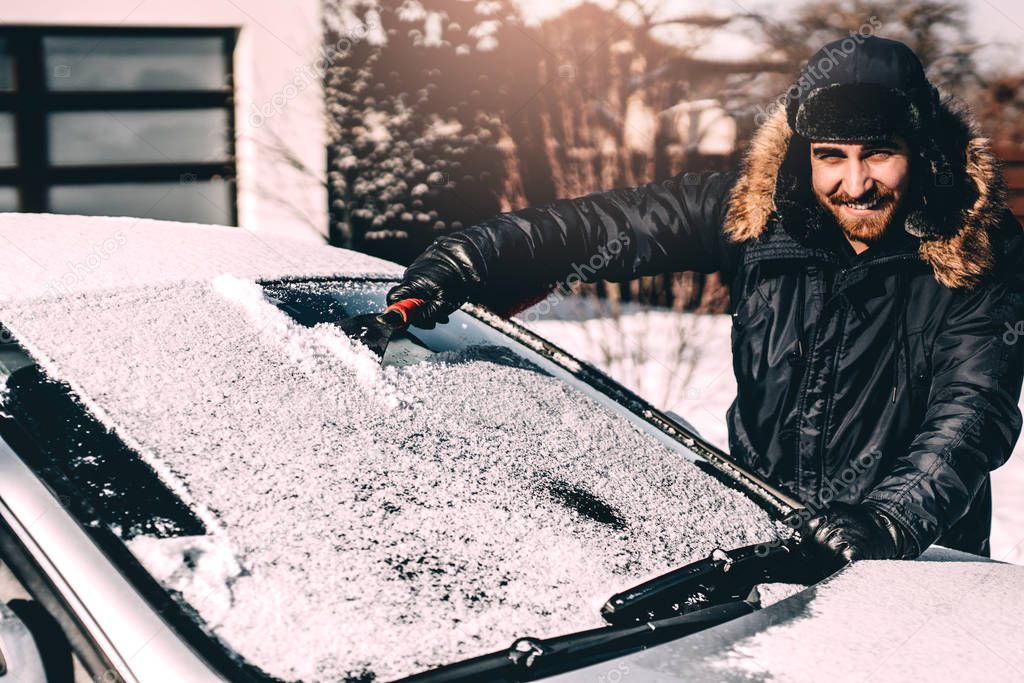 Attractive man smiling and cleaning snow off his car, working during winter and driving. 