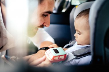 Father putting one year old baby in child car seat and smiling. Lifestyle, transportation concept clipart