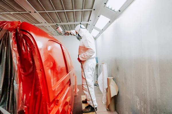 Travailleur peignant une voiture rouge dans une boîte de peinture spéciale, portant un costume blanc et gea de protection — Photo