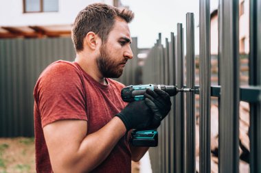 İnşaat işçisi elektrikli tornavida kullanıyor ve çitlere metal elementler yerleştiriyor.