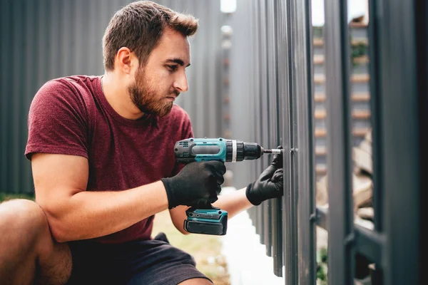 Blanke bouwman werkt met accuschroevendraaier op metalen hek — Stockfoto