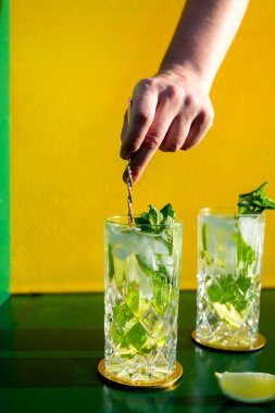 Fresh mojito alcoholic cocktail with rum, lime, mint and ice in a top shot glass shot with hard lights  and a hand stirring it with ice clipart