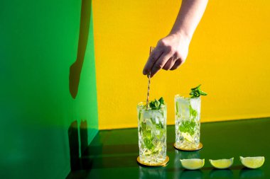 Mojito alkollü kokteyl ve barmen eli renkli arka planda buz karıştırıyor. Yeni modern kakaolar içki tasarımı konsepti