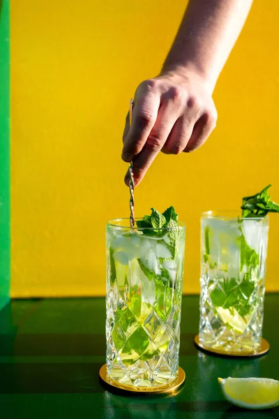 Rom, limon, nane ve buzlu taze mojito kokteyli. Sert ışıklarla çekilmiş bardakta. Bir el buzla karıştırıyor.