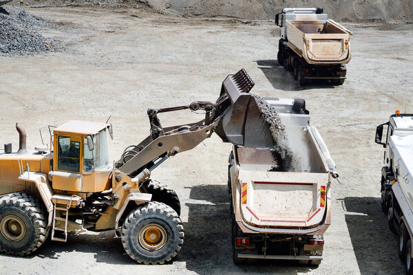 wheel loader with scoop working on construction site and loading gravel on dumper trucks. Heavy duty bulldozer working