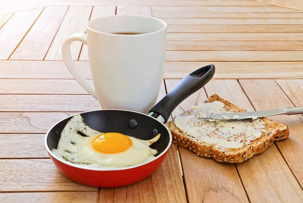 Close up of fried egg in pot, toast, and mug of coffee on wooden table — Stock Photo, Image