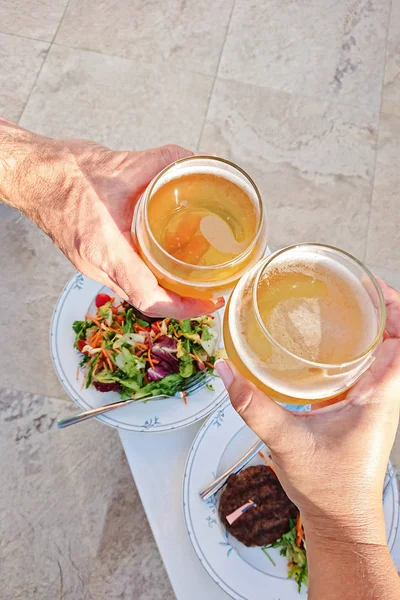 Manos sosteniendo vasos con cerveza sobre la mesa con platos de comida, vista superior . — Foto de Stock