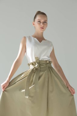 Stüdyoda poz veren kız. O, elinde geniş yeşil bir etek tutuyor ve düz görünüyor. Doğal makyaj. Belinde geniş kemer var..