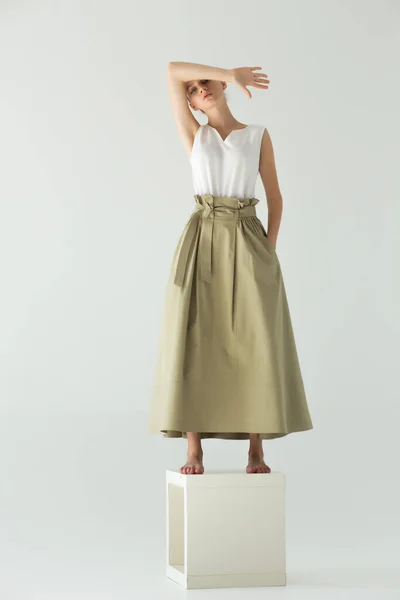 女模特光着脚站在工作室的一个立方体上 背景很浅 长裙和白色夏季T恤 胳膊弯了 盖住了她的额头 — 图库照片