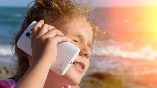 一个漂亮的小女孩用智能手机说话 微笑着 在阳光下斜视着大海的背景 日落时分 — 图库照片
