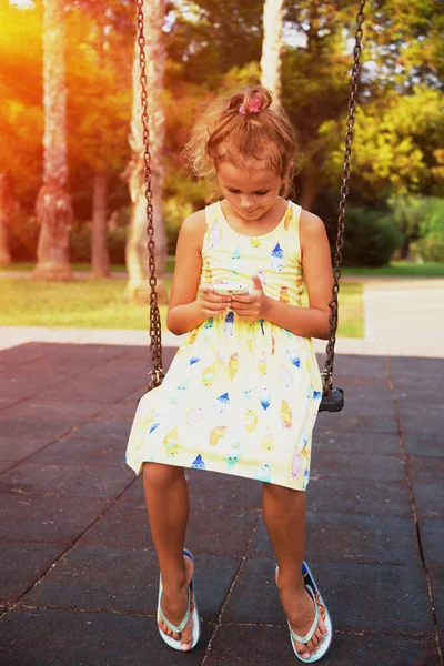 漂亮的小女孩在公园里的小孩游乐场里玩着智能手机 坐在链条上摇摆不定 她的注意力都集中在智能手机上了日落时间 美丽的阳光 — 图库照片