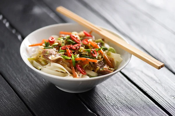 Traditional Asian noodles in minimalist style. Healthy nutritious meal of udon noodles, meat, vegetables, chili and spring onion in white bowl with chopsticks on black background copyspace