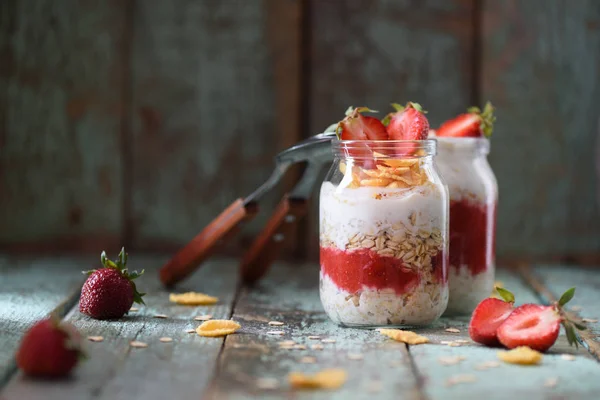 Healthy gluten free homemade breakfast. Overnight oats with strawberry and cornflakes in glass jars with wooden spoons on shabby blue background