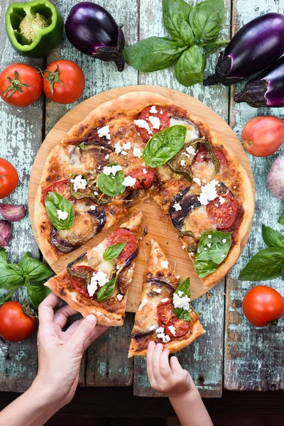 Tasty homemade rustic pizza. Woman and baby hands reaching for pieces of eggplant pizza with ricotta and basil top view