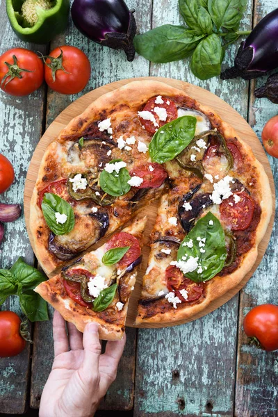 Healthy vegetarian pizza. Woman hand holding piece of aubergine pizza with ricotta, tomato, bell pepper and basil overhead view