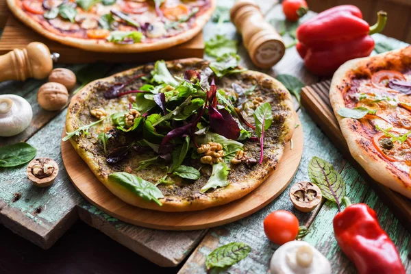 Homemade vegetarian pizzas. Thin crust rustic pizzas with tomatoes, mushrooms, pesto sause, leafy greens and nuts served with raw ingredients on shabby wooden background selective focus