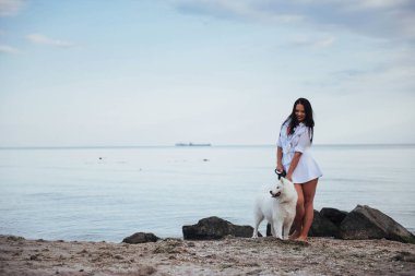 Köpeğiyle sahilde deniz kenarında yürüyen genç güzel kadın.