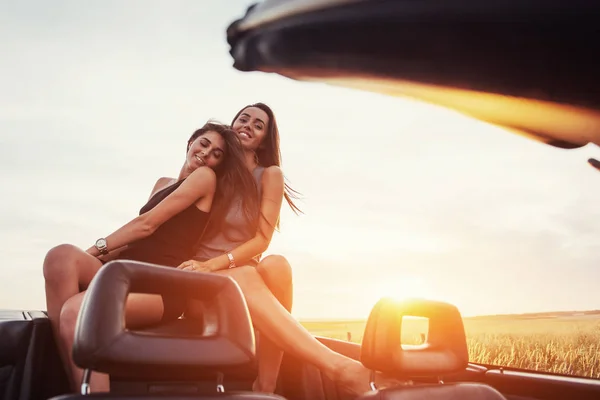 Young two women at a photo shoot. Girls gladly posing next to a black car against the sky on a fantastic sunset.