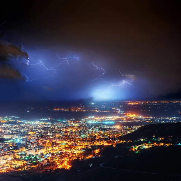 Night lights of the coastal city from the height. Lightning strike on the cloudy dark sky. Fantastic summer night scene.